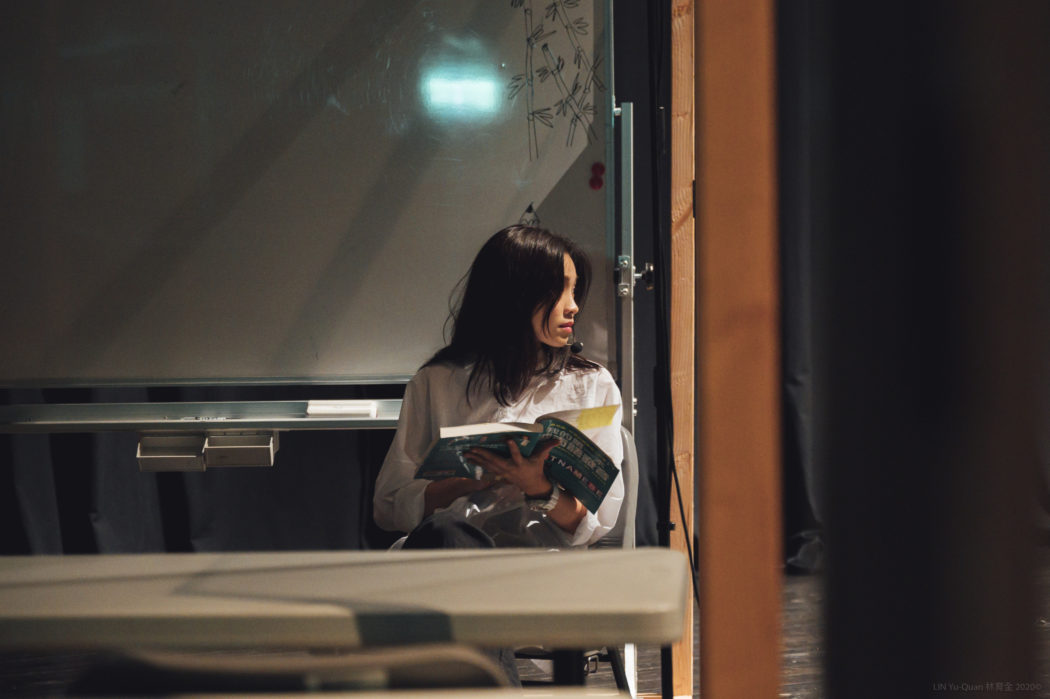 Eine vietnamesische Frau sitzt frontal an einem Tisch neben einem großen Fenster. Sie hält ein aufgeschlagenes Magazin in der Hand. sie schaut dabei nach rechts aus dem Fenster. Hinter ihr leuchtet ein grünes Exist-Schild. Der Fensterrahmen ist orange.
